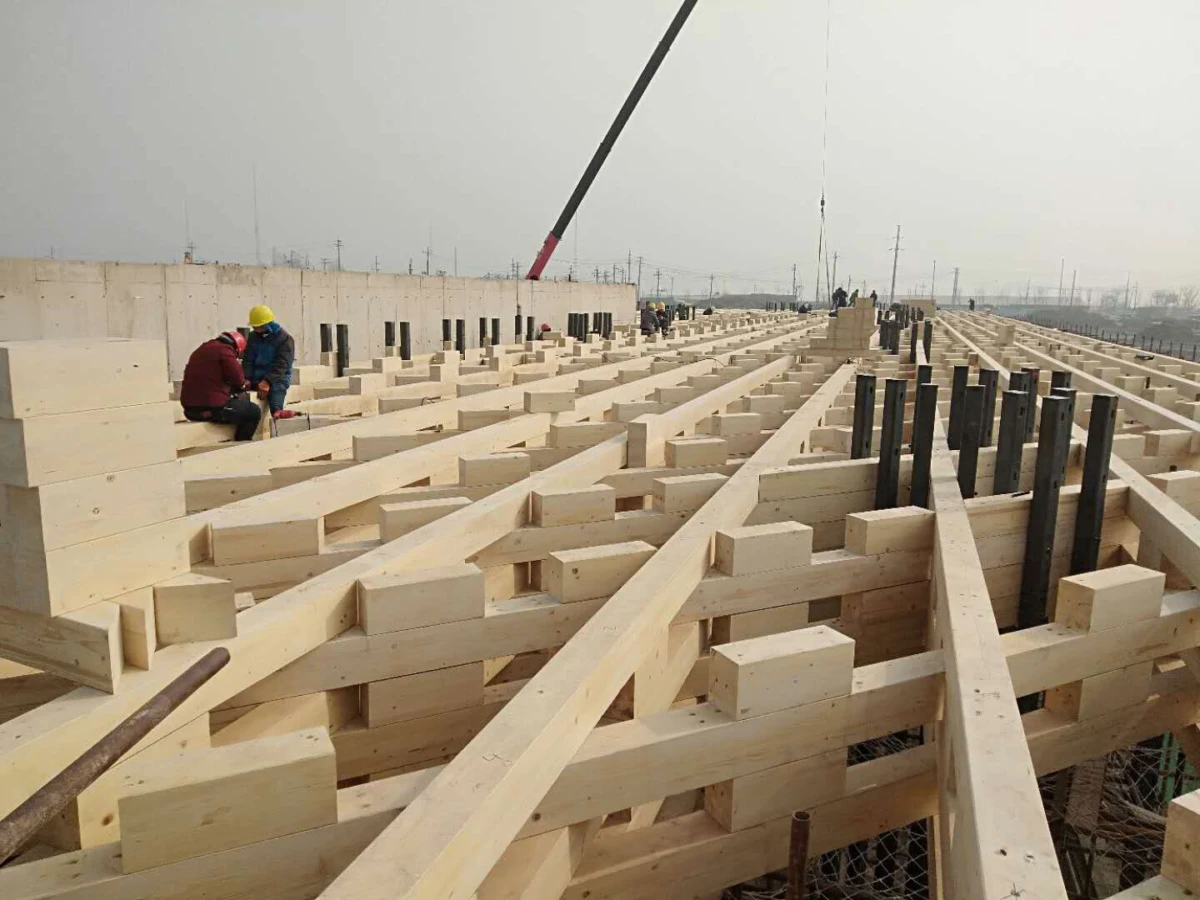 Construction workers in hard hats assemble a large wooden timber frame on an open construction site.