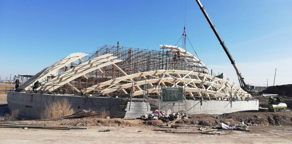 Desert construction site with a dome-shaped wooden lattice on a concrete foundation; crane lifts panels over a scaffolded frame.