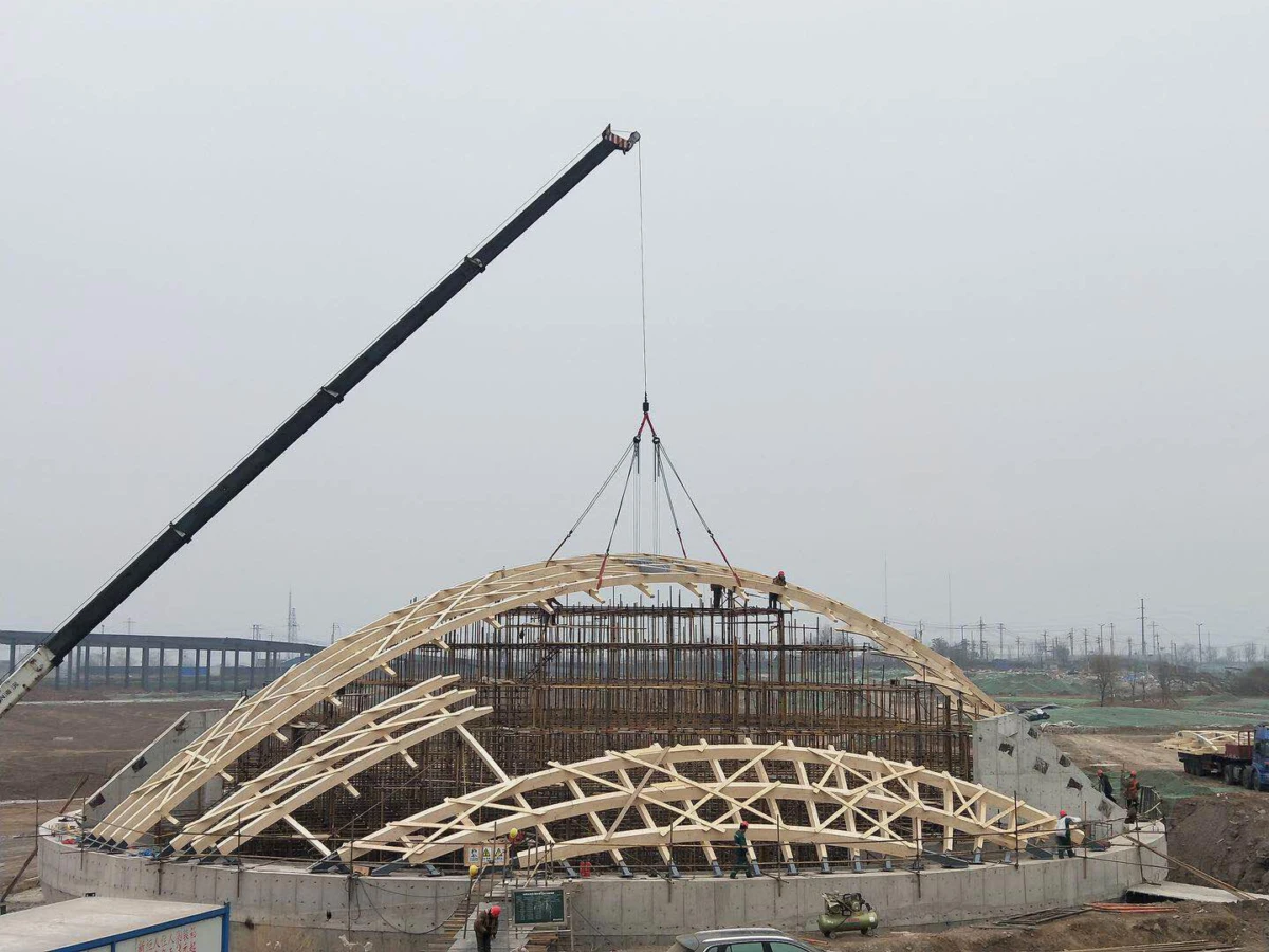 Crane lifts a geodesic wooden dome frame onto a circular concrete foundation at a construction site.