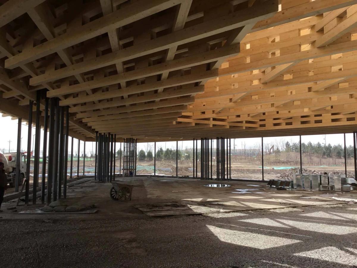 Interior of an under-construction building: wooden roof trusses, steel columns, open sides, construction debris on dirt floor.