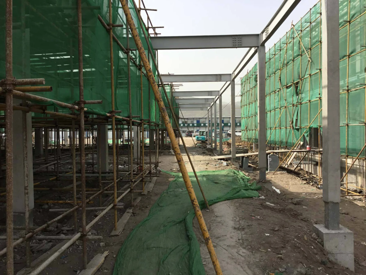 Outdoor construction site with metal scaffolding and green netting on both sides, concrete columns, and a green ground tarp along central aisle.