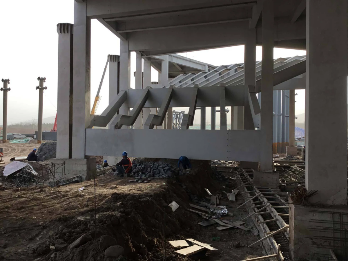 Construction workers assemble a large steel-girder framework beneath concrete columns at an outdoor construction site.