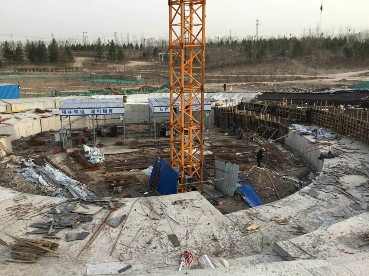 Construction site with an orange tower crane over a circular excavation pit; workers and scattered materials.