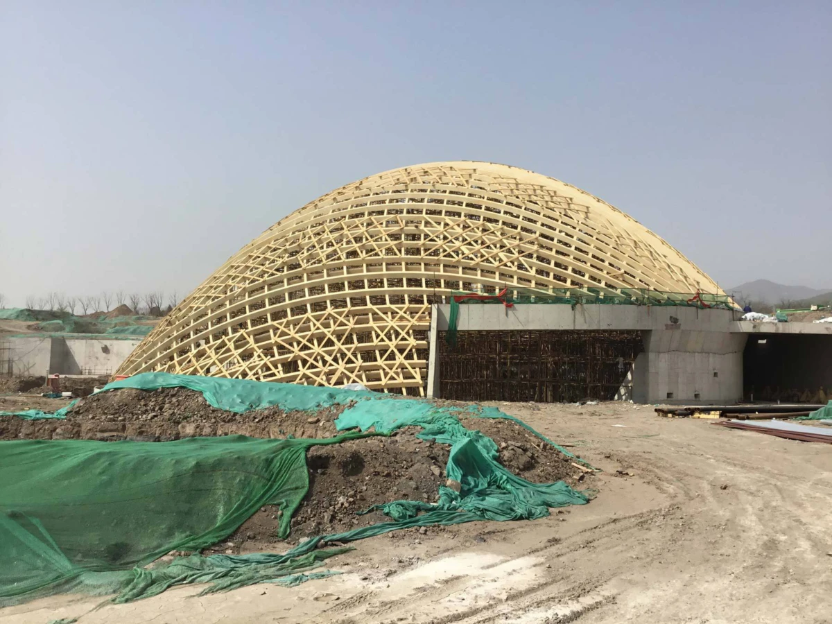 Geodesic wooden lattice dome under construction at a building site; green tarps and dirt piles surround the unfinished structure.