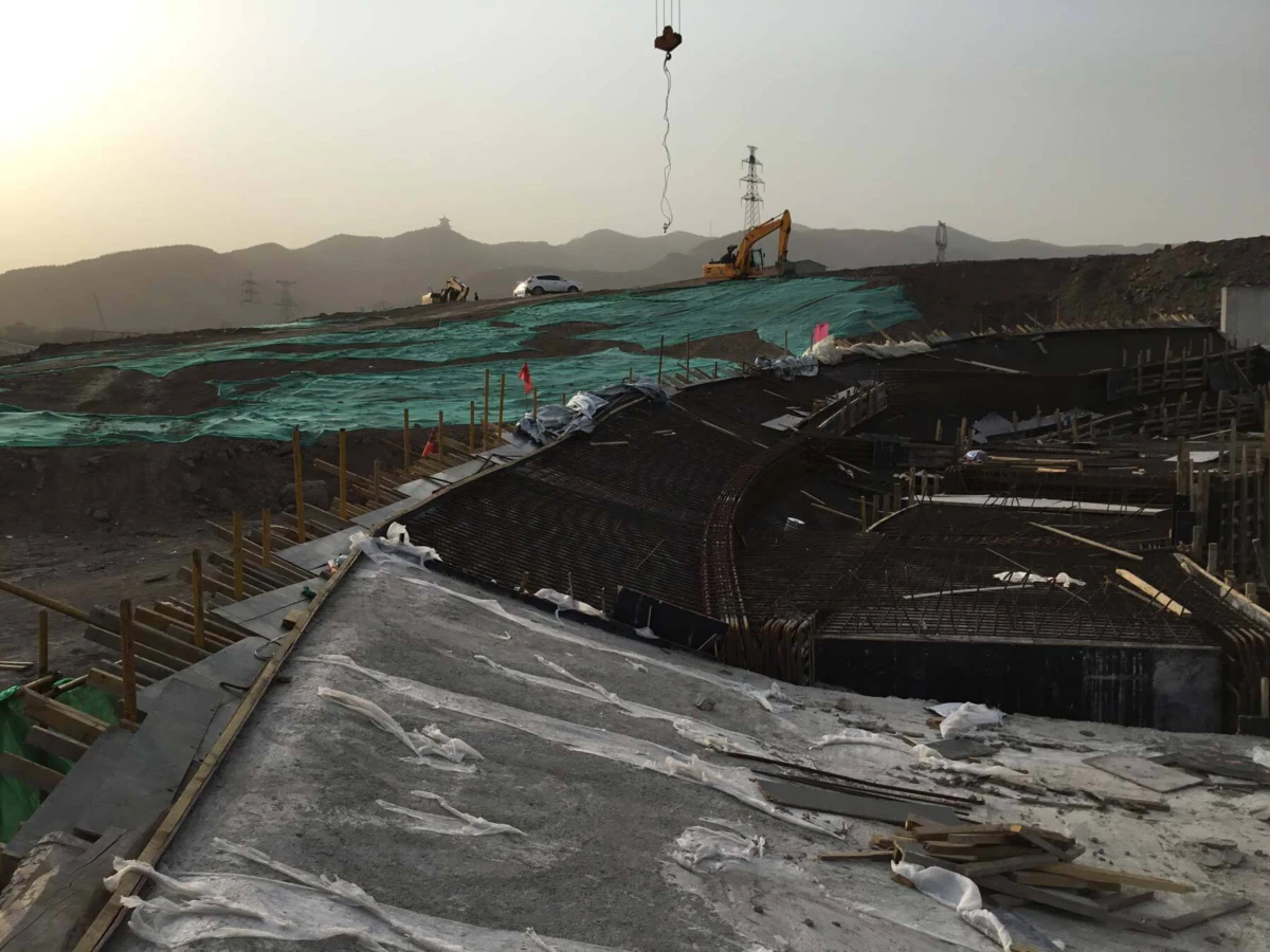 Construction site featuring a curved reinforced-concrete foundation with exposed rebar, scaffolding, and a yellow excavator on a hillside at sunset.