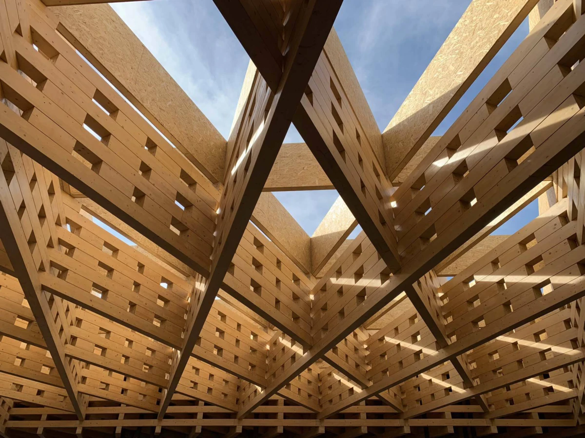 Under-construction wooden timber lattice framework; intersecting beams form geometric openings against a blue sky.