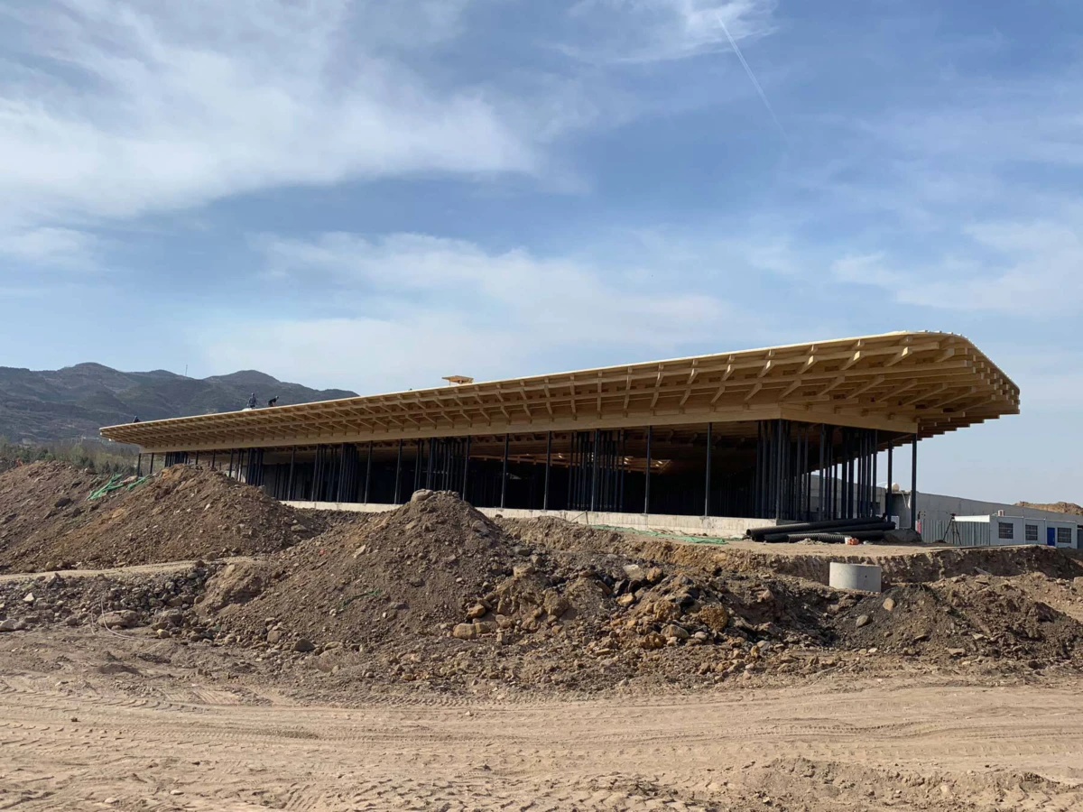 Construction site: a long building with a curved wooden roof supported by steel columns, dirt mounds in the foreground, hills in the distance.