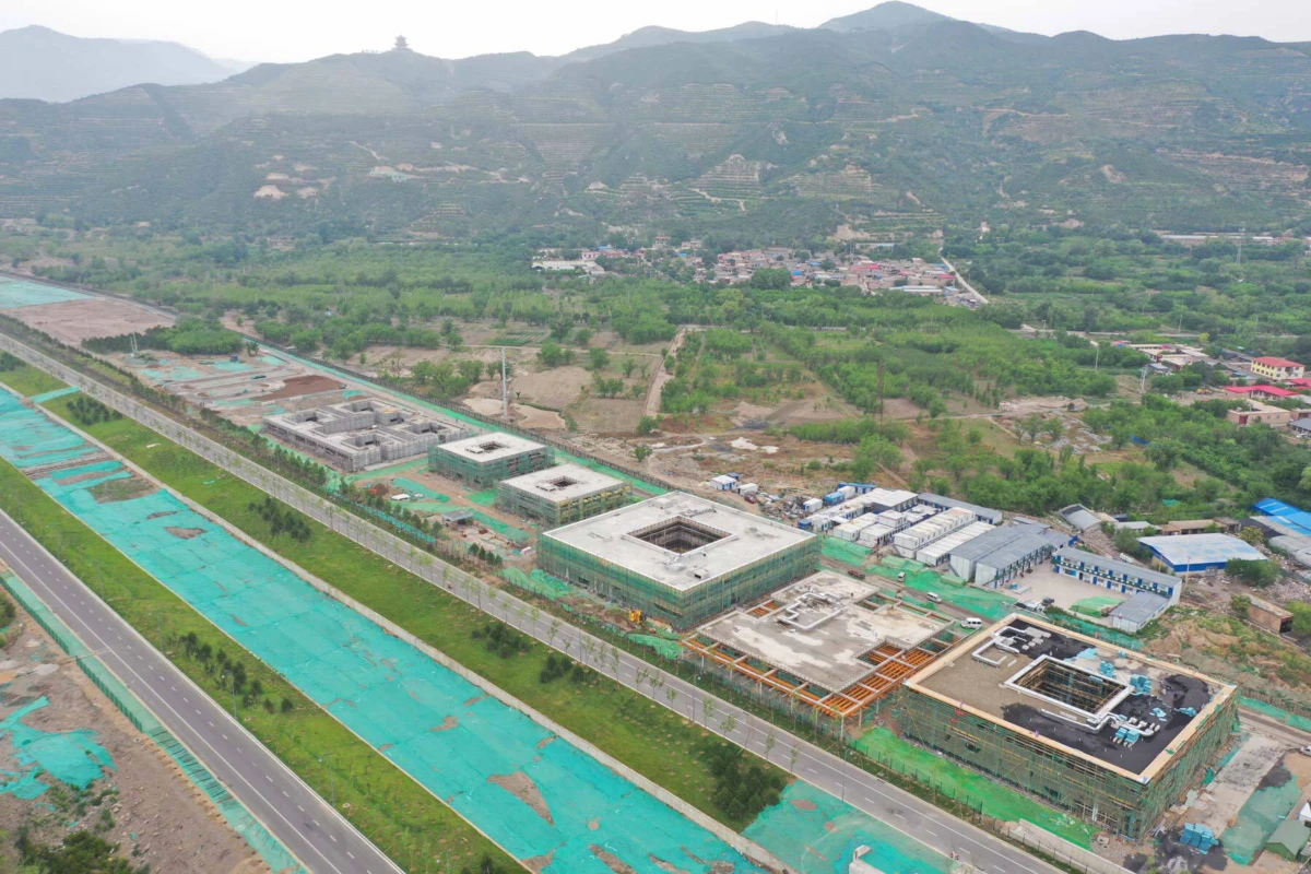 Aerial view of a construction site with square concrete buildings under green netting along a highway in a valley with distant hills.