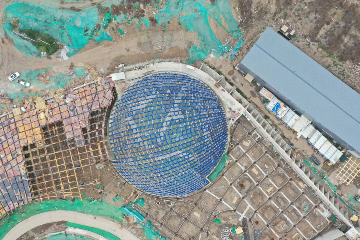 Aerial view of a construction site featuring a large blue geodesic dome under construction, with scaffolding and a long metal shed nearby.