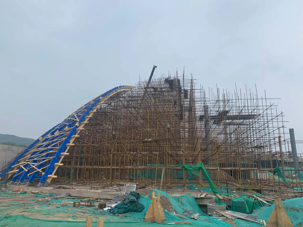 Large bamboo scaffolding surrounds a curved blue arch frame at a construction site with green tarps and debris