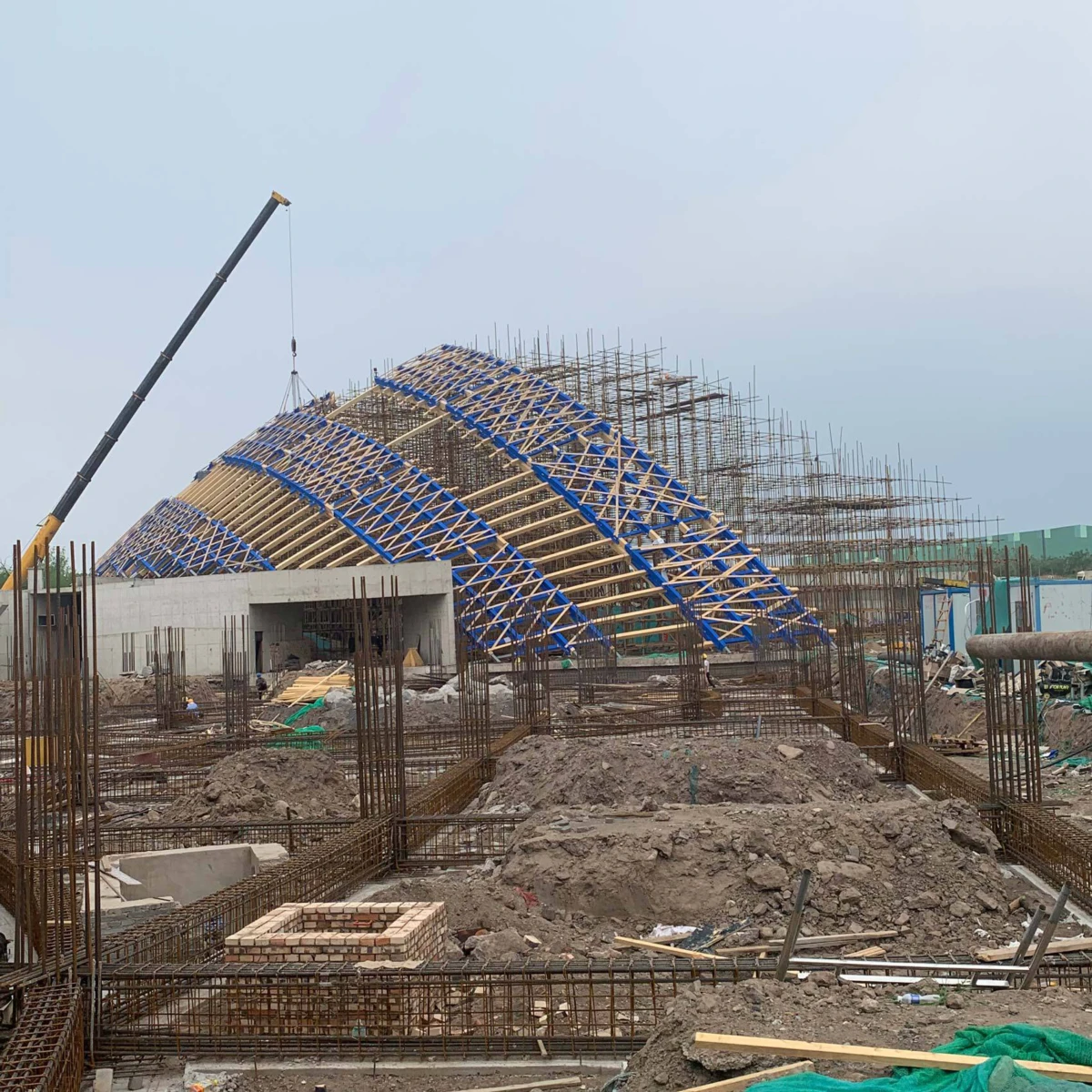 Geodesic lattice dome under construction with blue bracing at a crane-equipped building site.