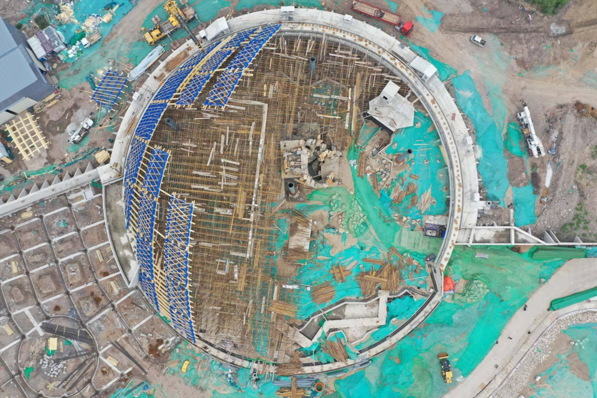 Aerial view of a circular concrete structure under construction, with extensive wooden formwork, steel rebar, and blue scaffolding around the ring.