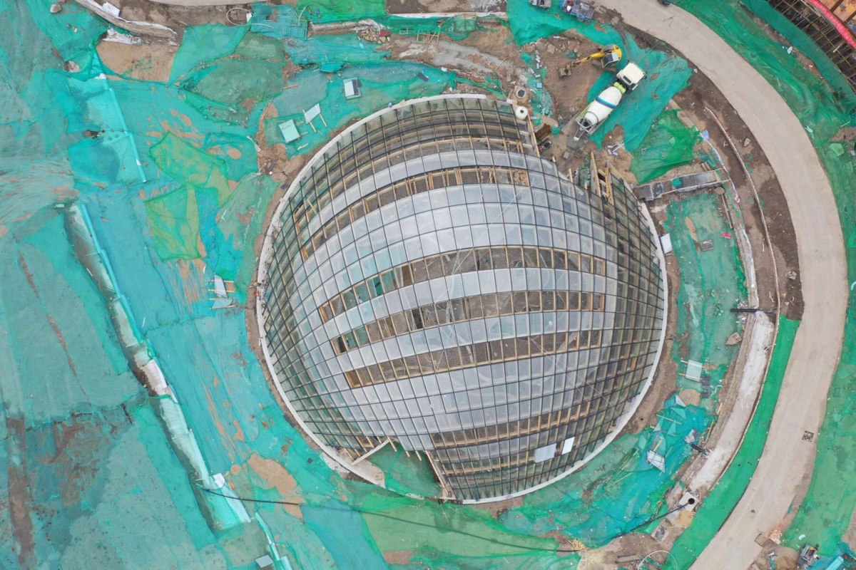 Geodesic glass dome under construction, encircled by green construction netting.