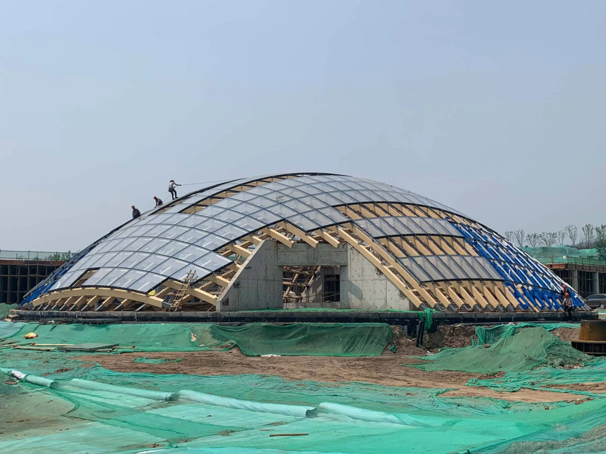 Geodesic glass dome under construction with wooden ribs; workers on top at an outdoor building site.