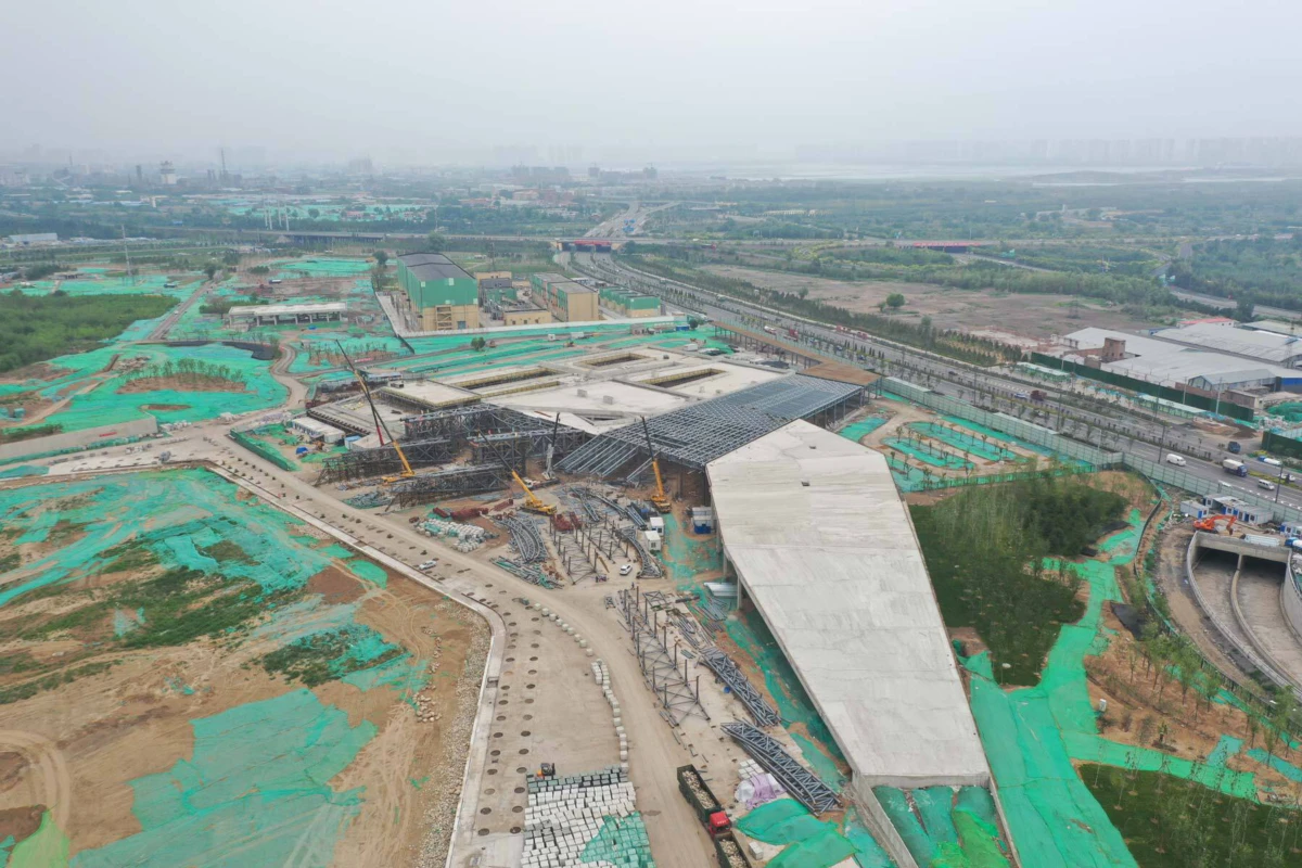 Aerial view of a large construction site with cranes and a curved concrete ramp, surrounded by green safety netting and roads.