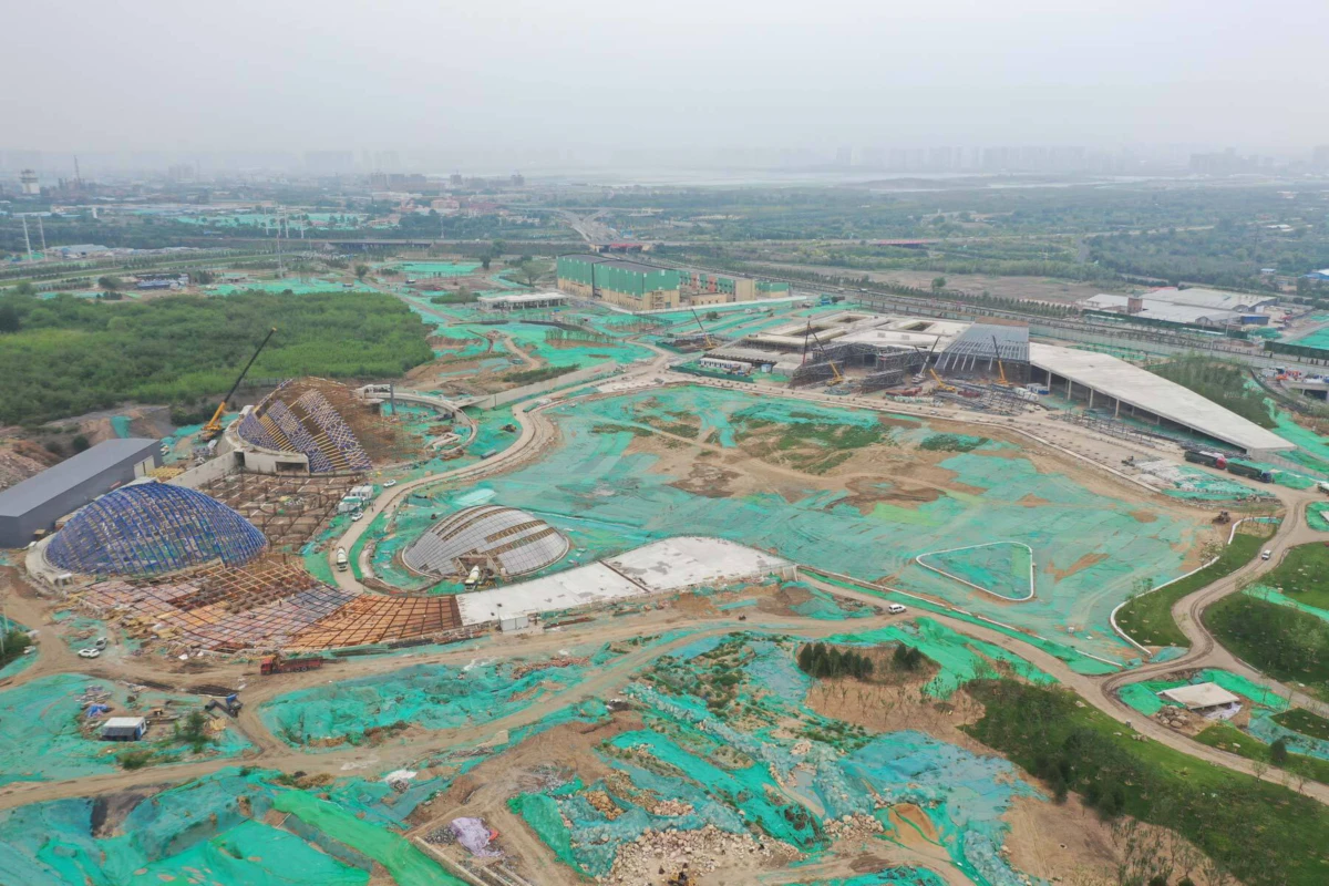 Construction site with several geodesic domes under construction, green netting over the ground, distant city skyline.