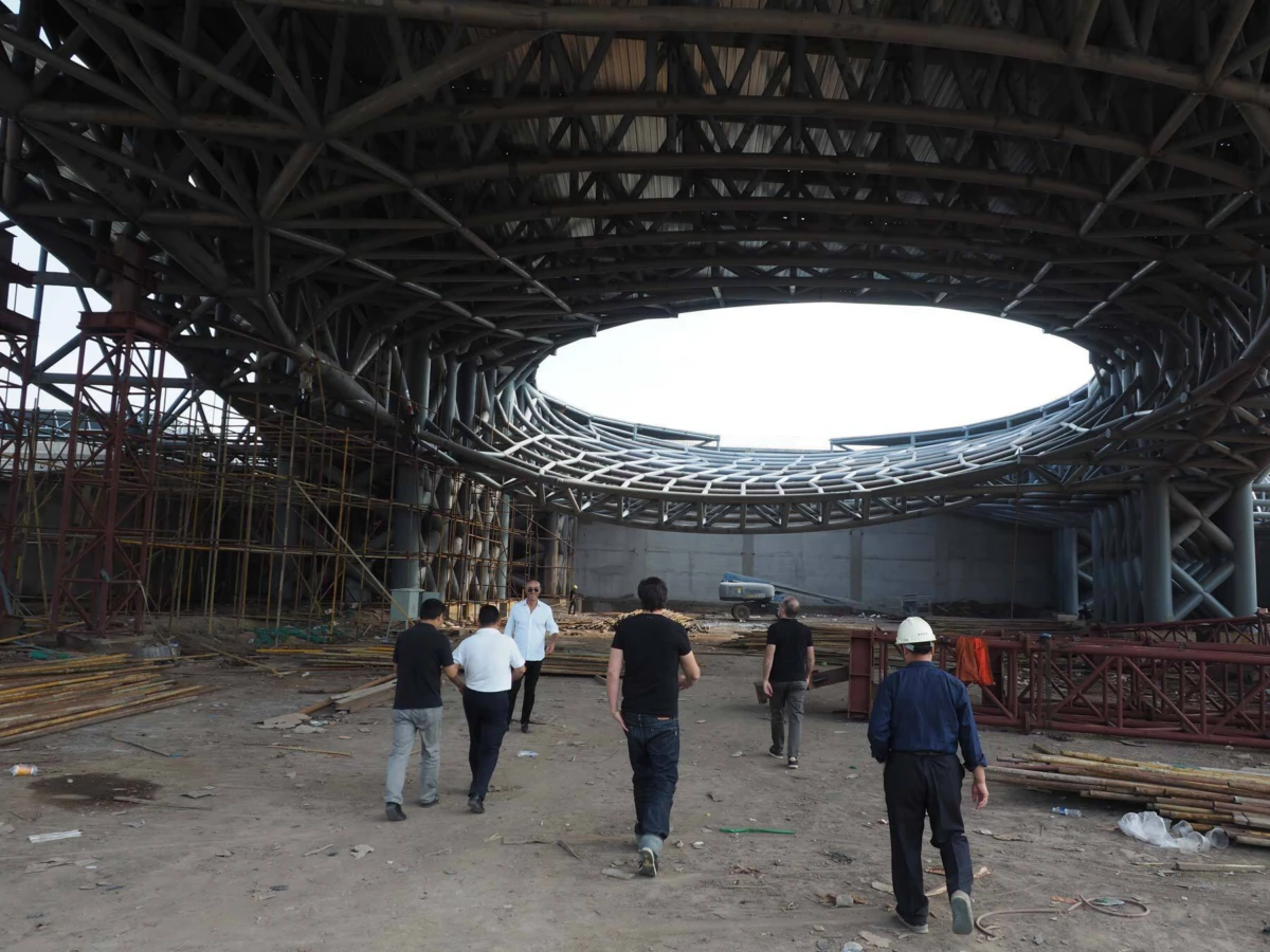 Construction crew walking inside a circular steel-lattice dome under construction; scaffolding and materials on the dirt ground.
