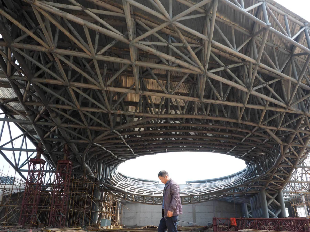 Person in a gray jacket walks beneath a complex curved steel lattice canopy at a construction site.
