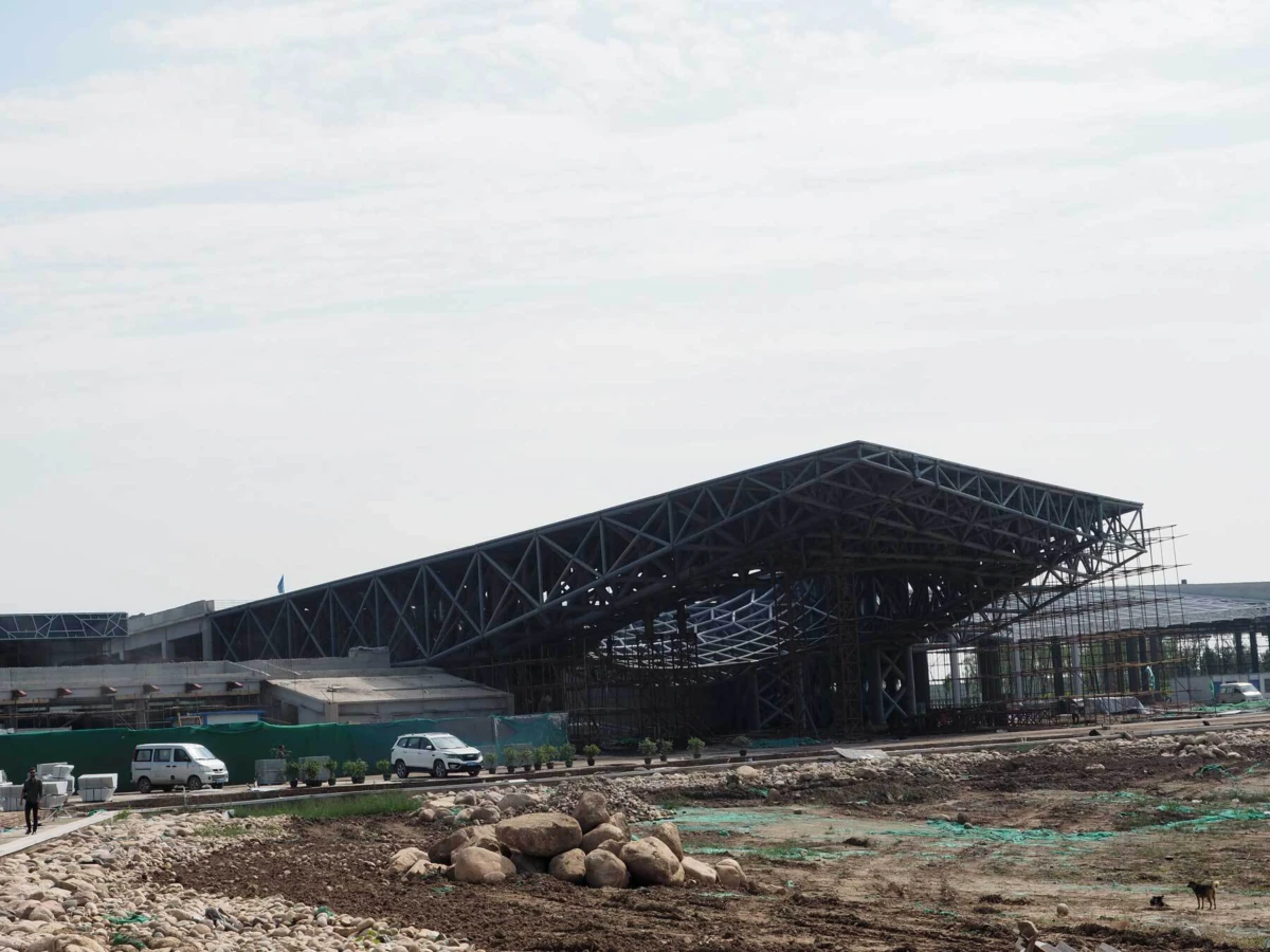 Large steel lattice roof structure under construction at an industrial site; rocky foreground with white vans nearby.