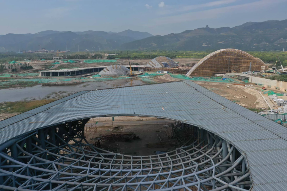 Circular steel lattice framework of a building under construction, with curved domes and distant mountains.