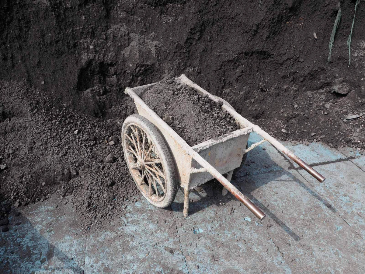 Rusty wheelbarrow filled with soil rests beside a large dirt mound on a dusty concrete surface.