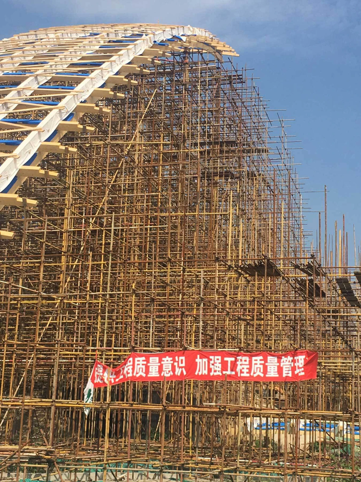 Bamboo and steel scaffolding forming a curved arch at a construction site; a red banner with Chinese text hangs in front.