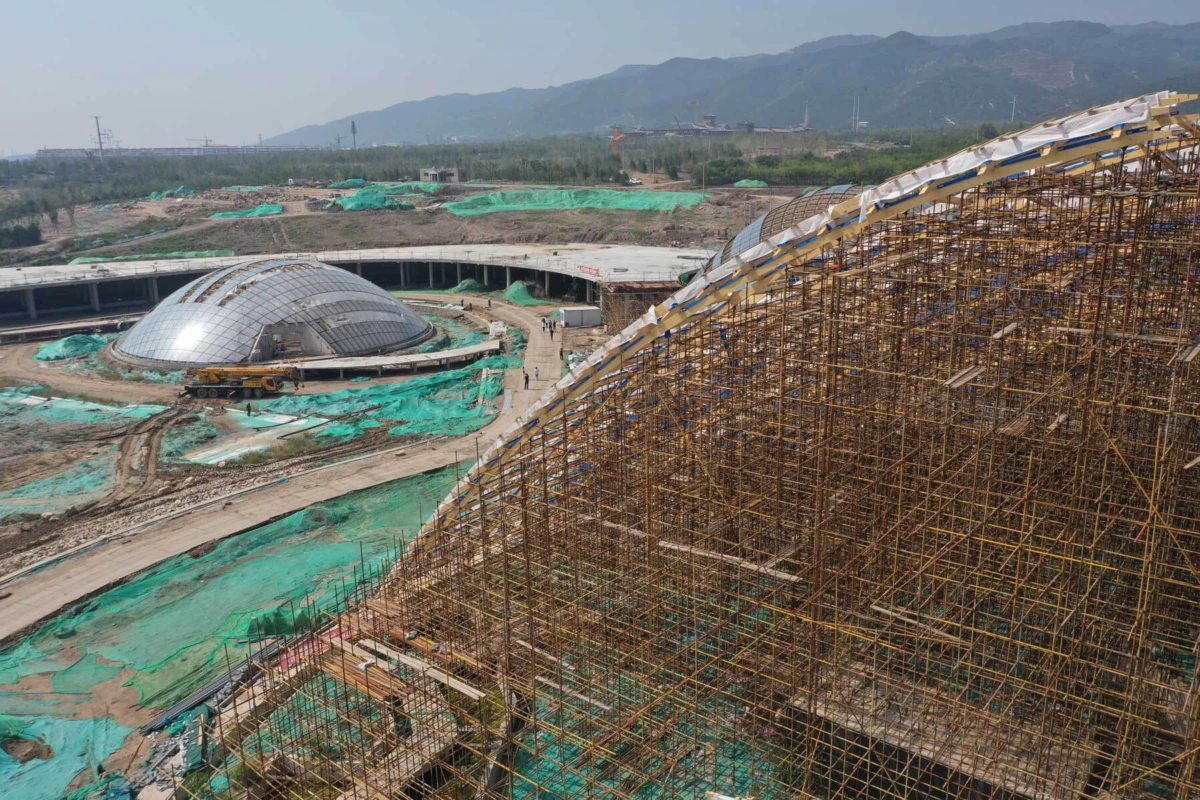 Large construction site with dense scaffolding around a curved-roof structure and geodesic domes; green safety netting and distant mountains.