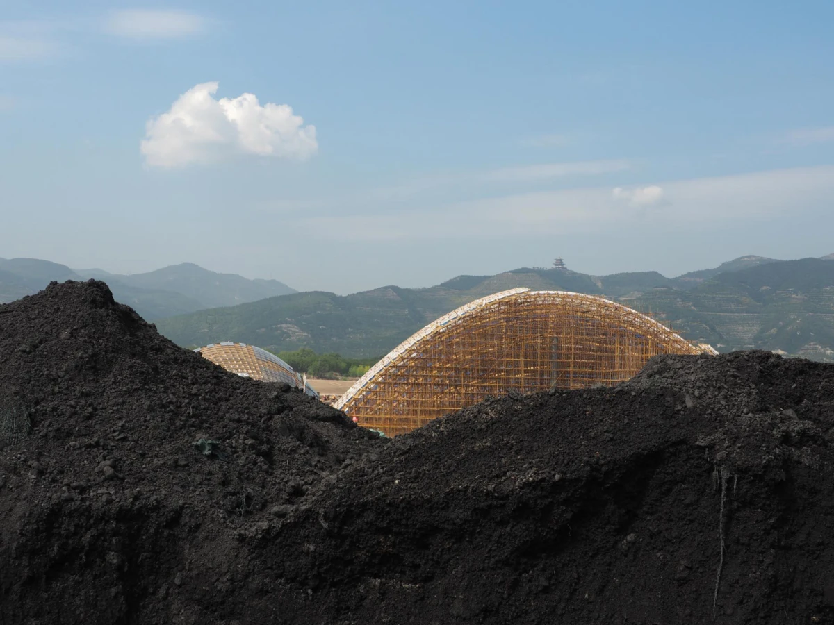 Wooden lattice arch framework under construction, dark soil piles in the foreground, with distant mountains under a blue sky.