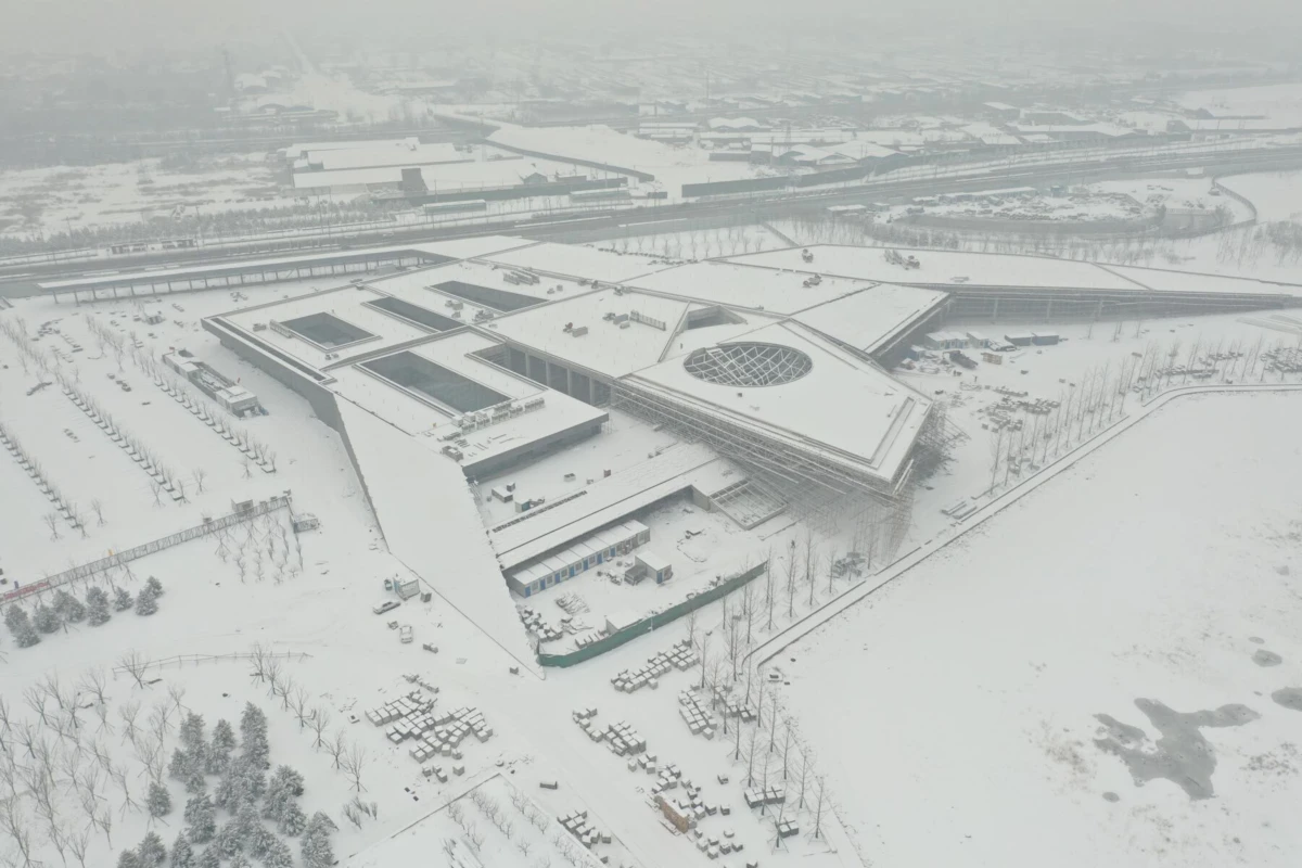 Aerial view of a large modern building complex with a circular glass dome, snow-covered, surrounded by roads and leafless trees.