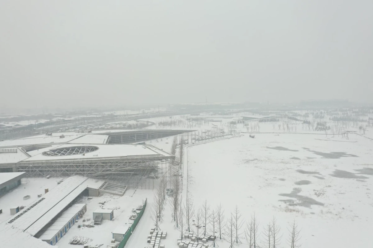 Aerial view of a snow-covered urban complex with a large modern terminal featuring a circular glass roof, bare trees, and frozen fields.