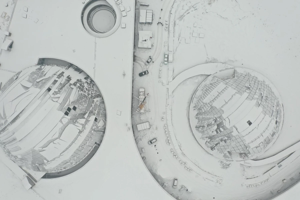 Aerial view of two large geodesic glass domes in a snow-covered landscape, with a road and construction vehicles between them.
