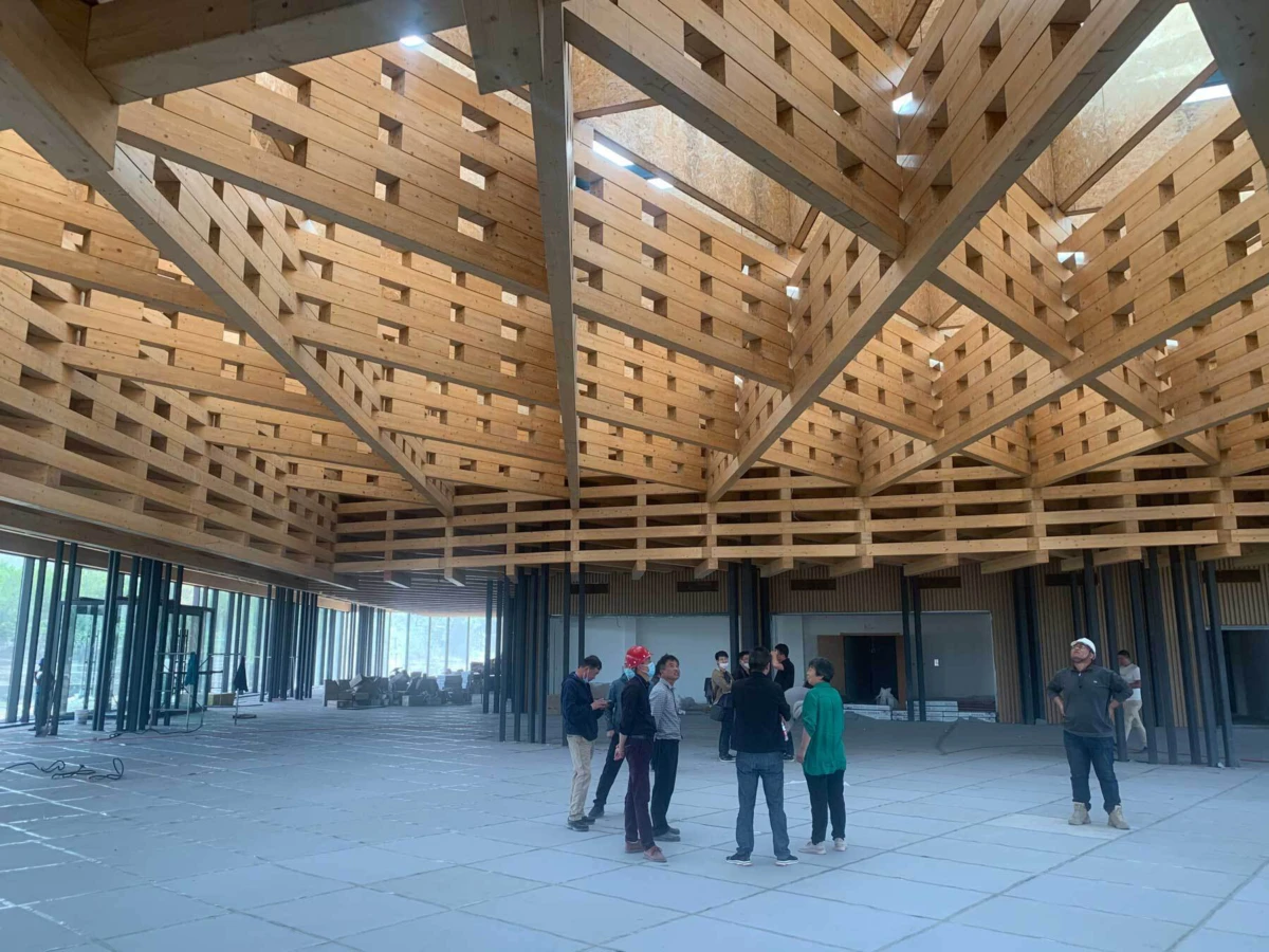 Group of construction workers in hard hats standing in a circle inside a large unfinished hall with exposed timber roof trusses.