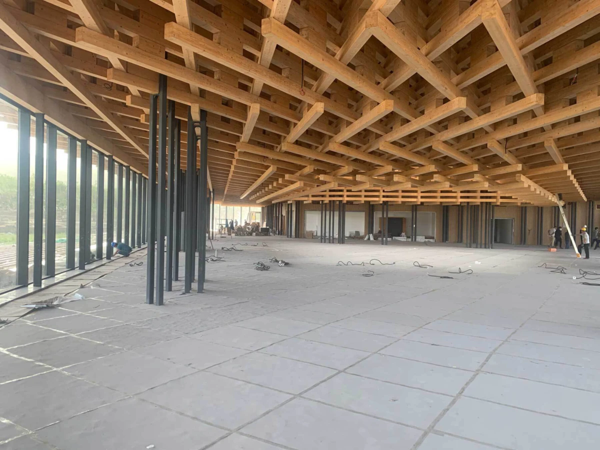 Interior of a large building under construction with exposed wooden roof trusses and steel columns; construction workers in the background.