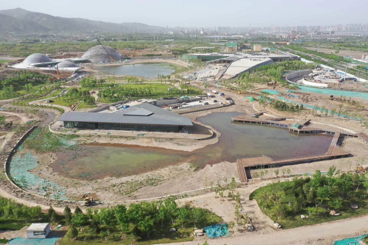 A construction-site eco-park with a pond, wooden boardwalks, and a low angular-roof building; geodesic domes and distant city skyline.