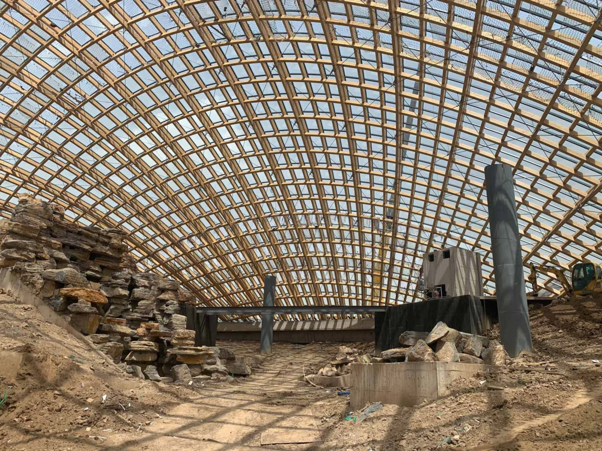 Inside a large construction site under an arched wooden lattice roof, with stone piles, dirt floor, and construction equipment.