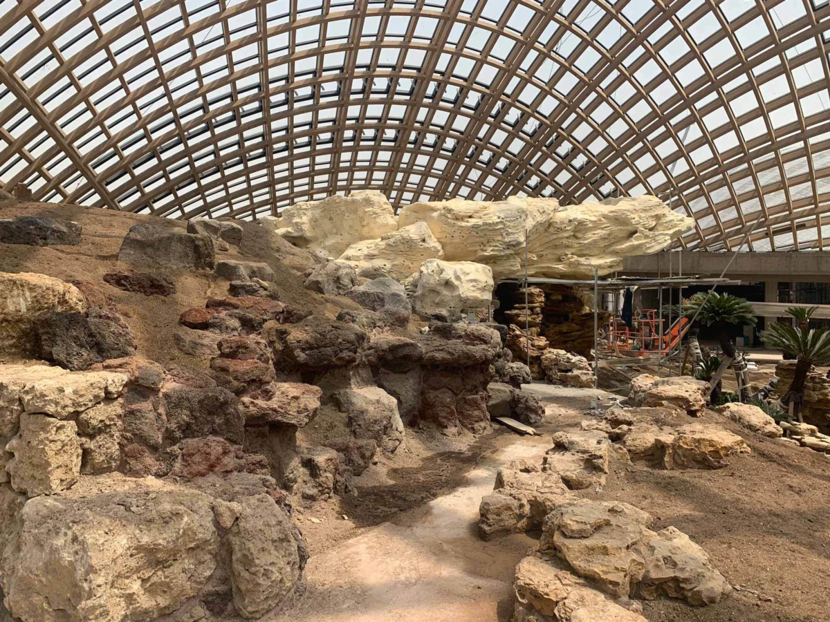 Rocky ruin exhibit under glass-dome ceiling; indoor museum scene with scaffolding and tropical plants.