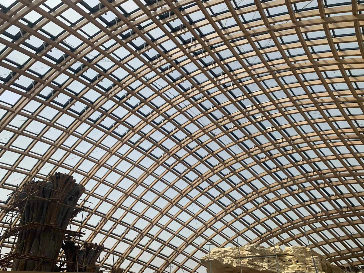 Interior with a curved wooden lattice roof and glass panels; a bundle of wooden logs bound with wire in the foreground beside a rocky formation.