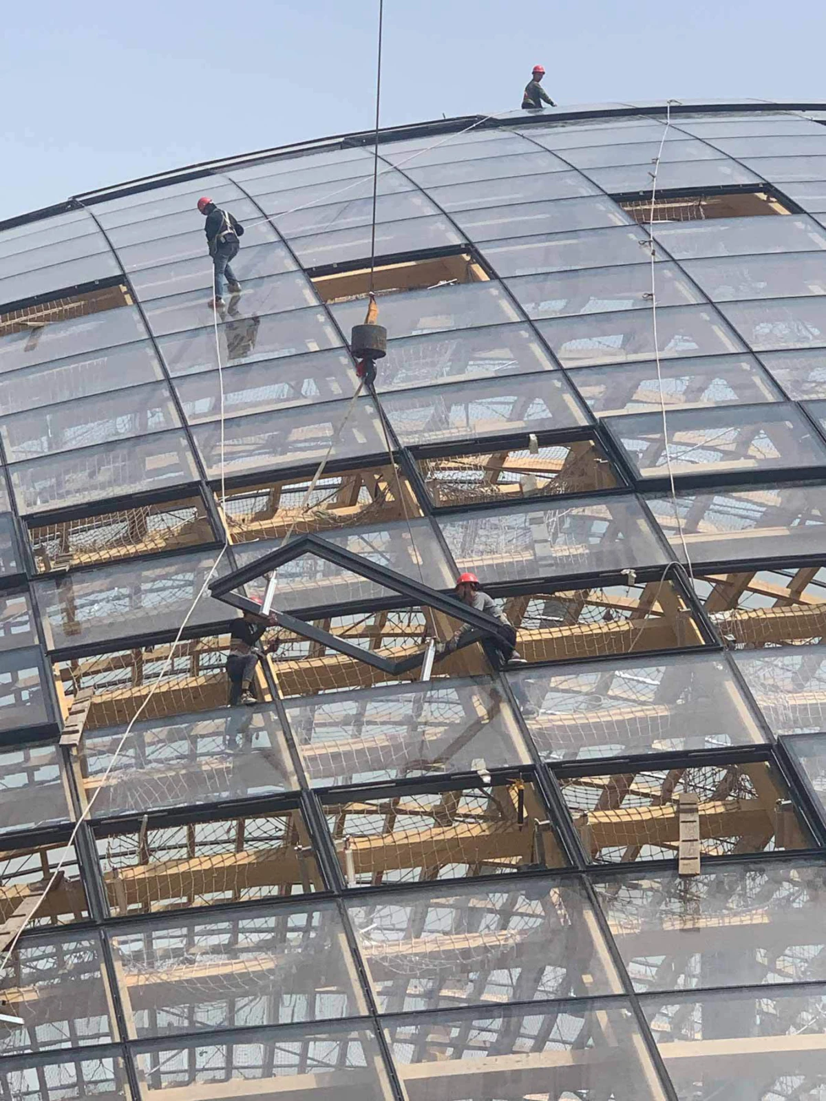Four construction workers in red helmets install a metal beam on a curved glass dome, secured by ropes.
