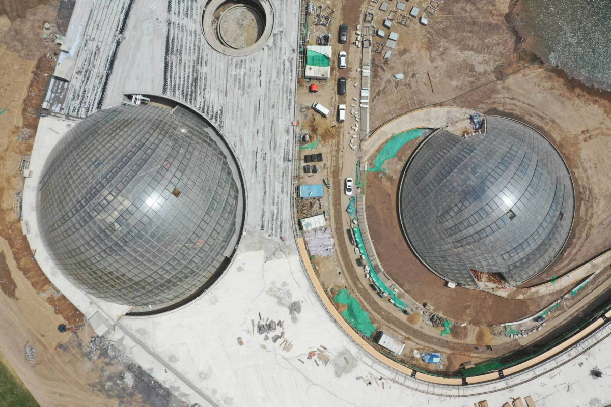 Aerial view of two large glass domes under construction, with dirt ground, circular bases, and construction vehicles.