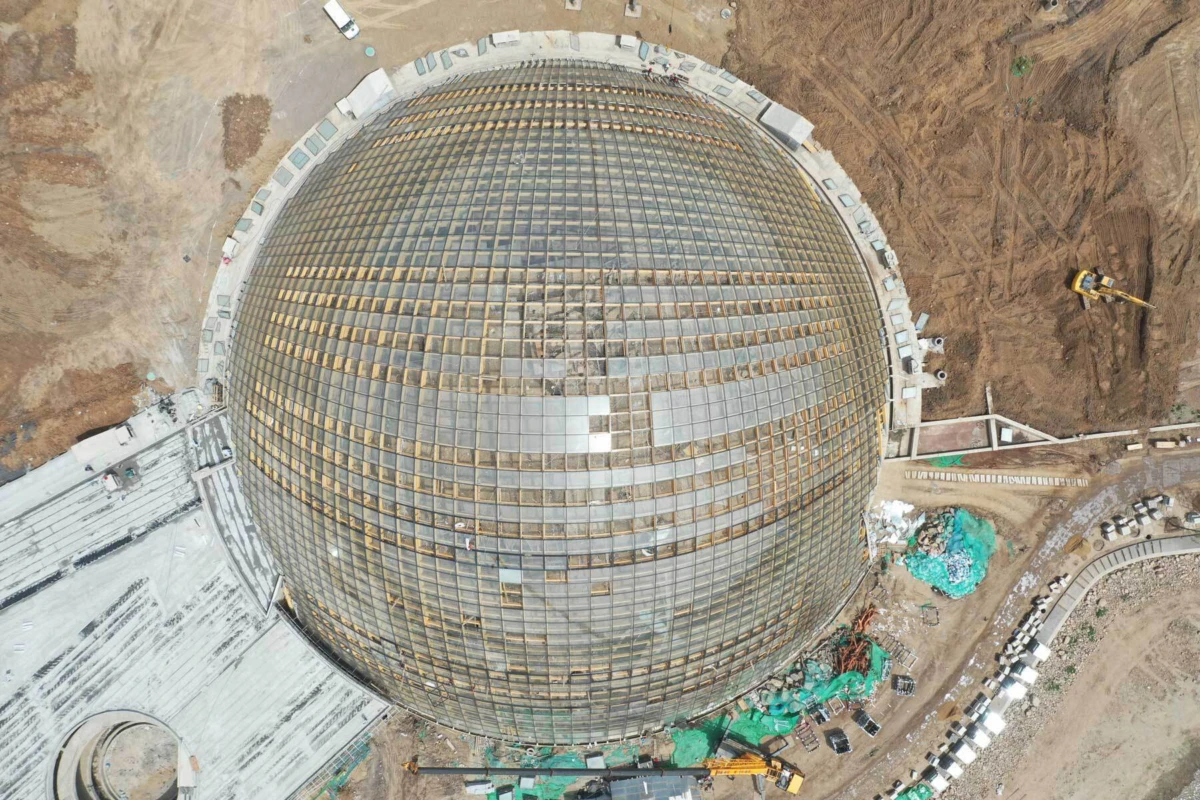 Aerial view of a large geodesic glass dome under construction at a muddy construction site with machinery.