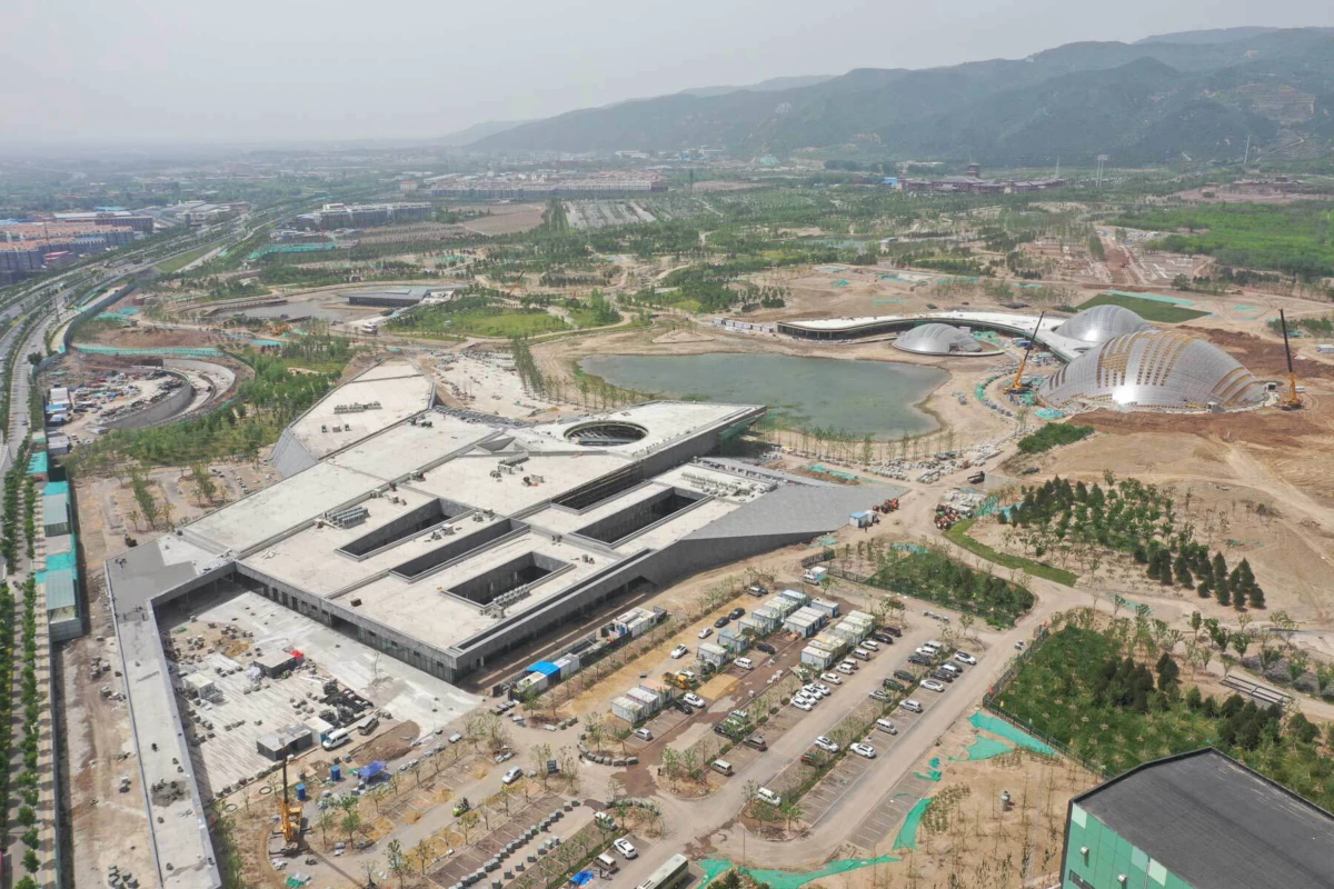 Aerial view of a large construction site with an L-shaped concrete building, two metallic domed pavilions, and a surrounding lake and hills.