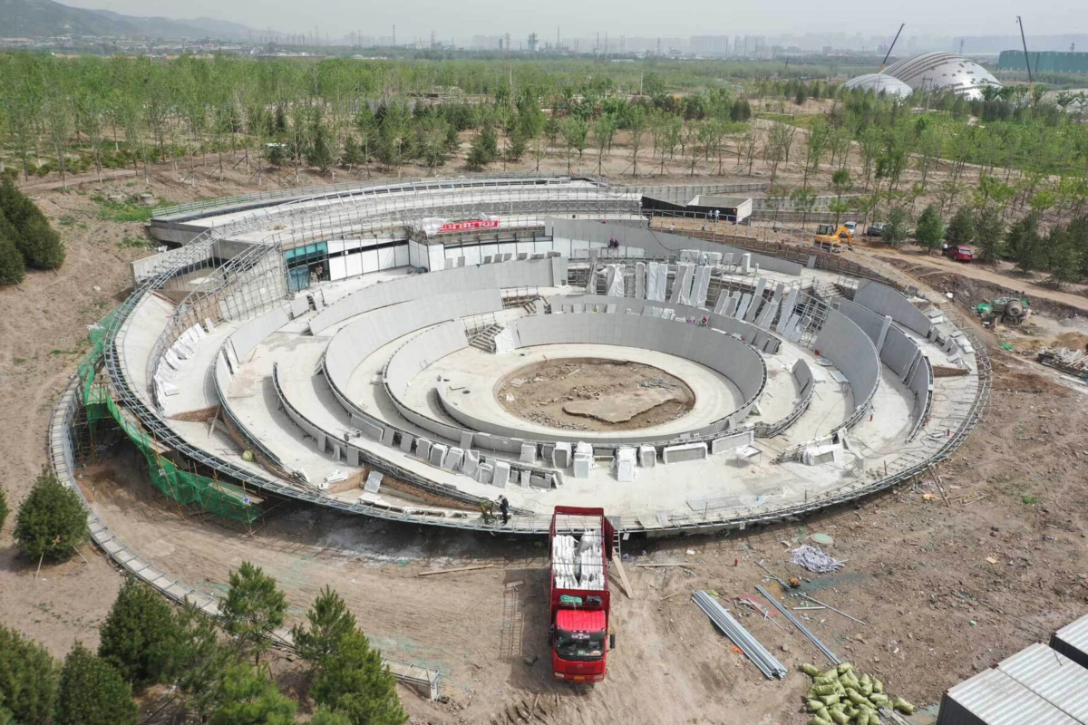 Circular concrete amphitheater under construction with concentric seating rings, on an outdoor site with a red truck nearby among trees.