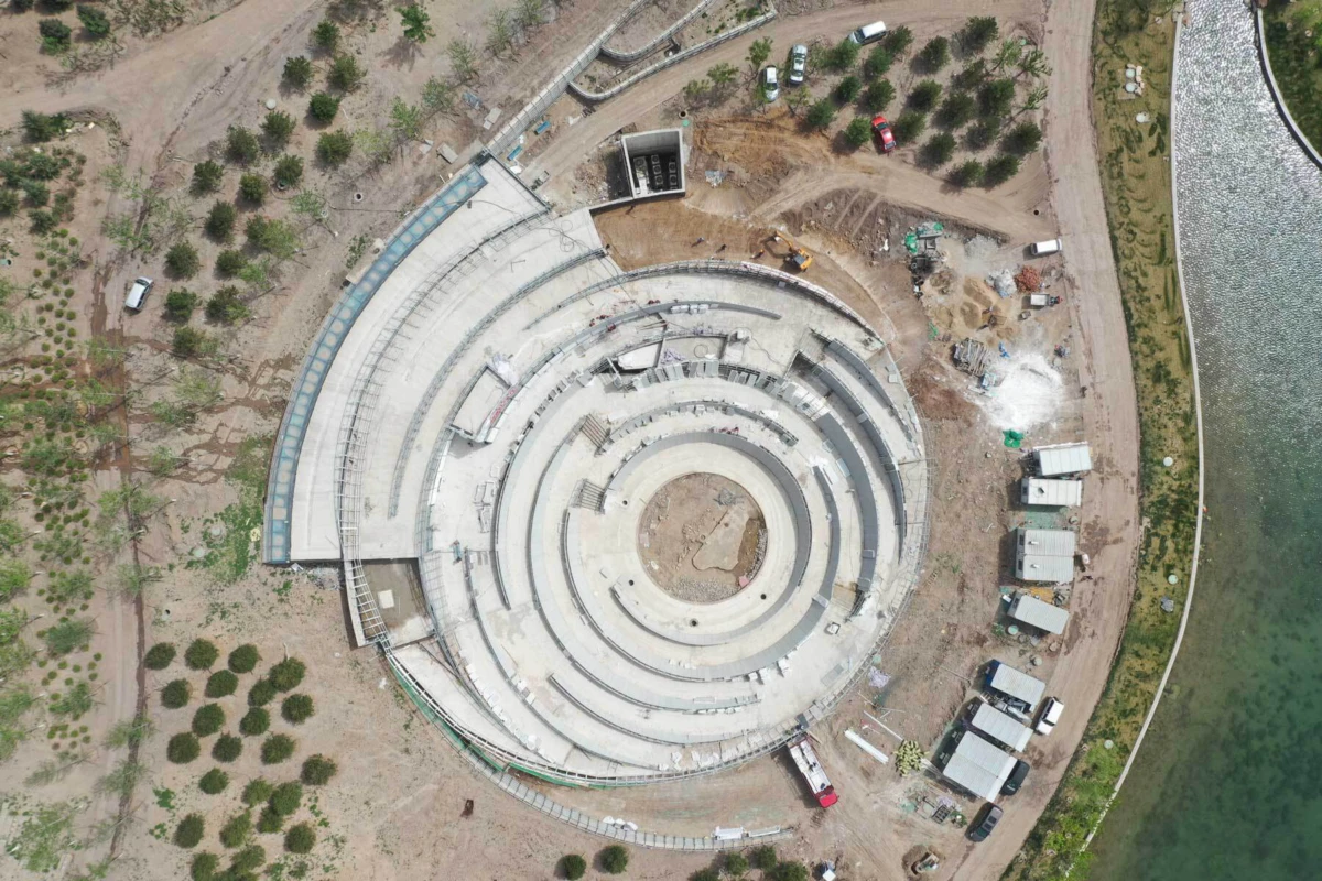 Aerial view of circular concrete amphitheater under construction along a shoreline, with construction machinery and trucks nearby.
