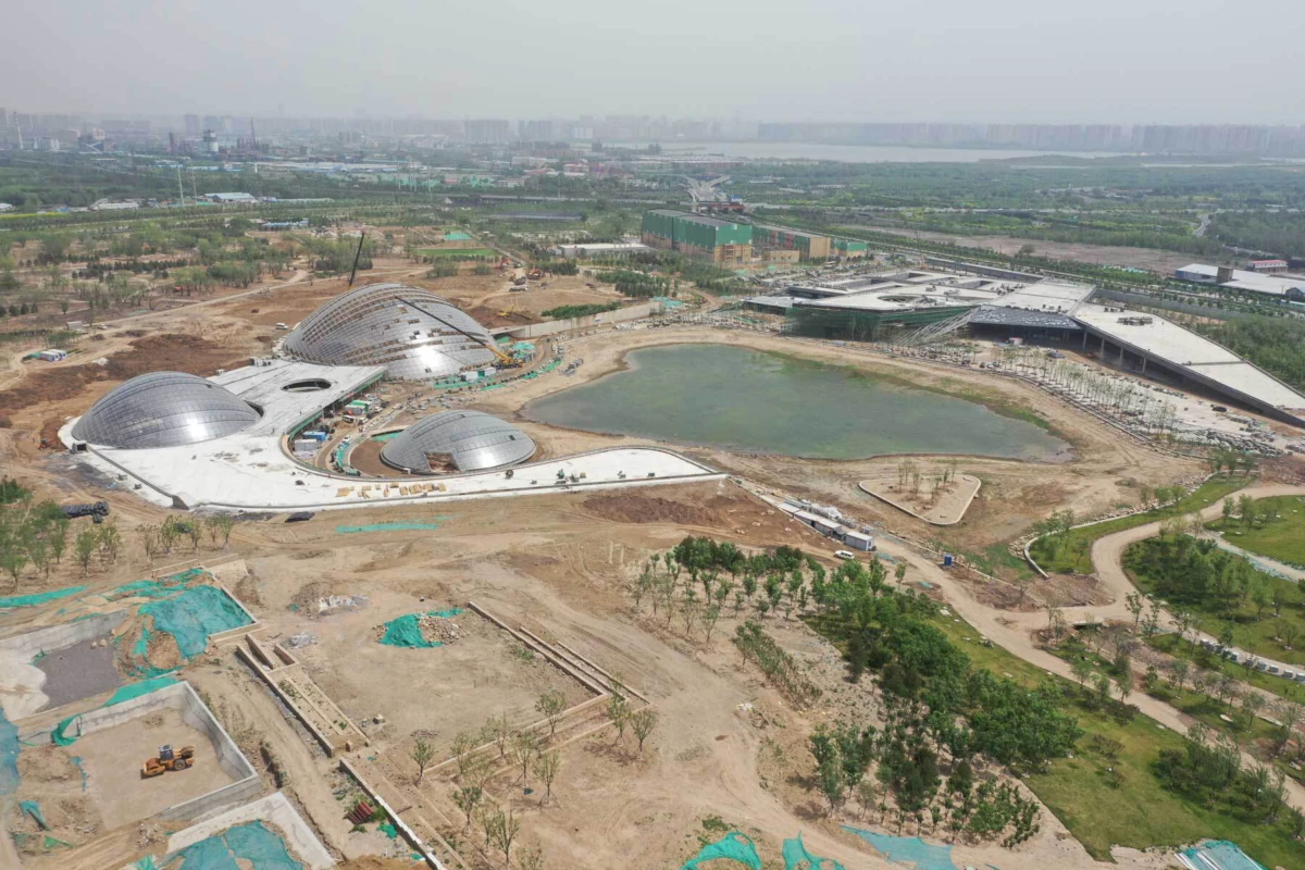 Aerial view of a construction site with dome-shaped buildings around a central pond; surrounding greenery and distant city skyline.
