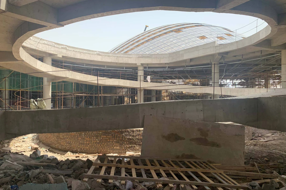 Under-construction circular spiral concrete ramp with scaffolding; construction workers on site, brick interior wall and glass dome overhead.