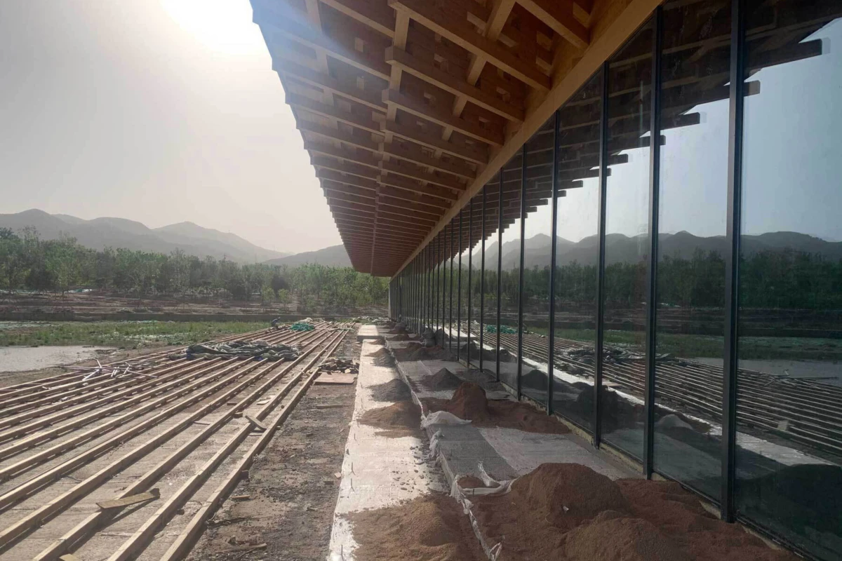 Construction in progress beside a glass-walled building; exposed wooden roof beams, metal pipes, and sand piles; distant hills.