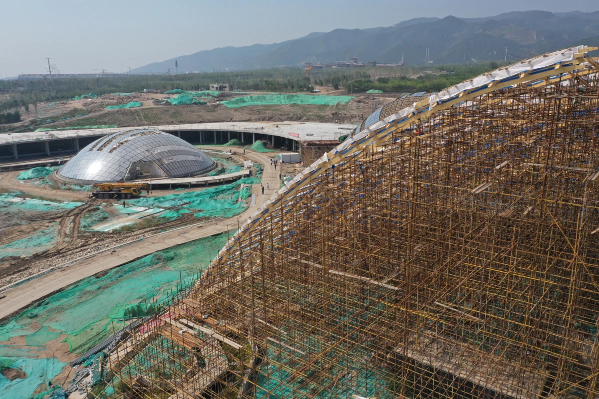 Construction site with a large bamboo scaffold on the right and two glass geodesic domes on the left, with green tarps and distant mountains.