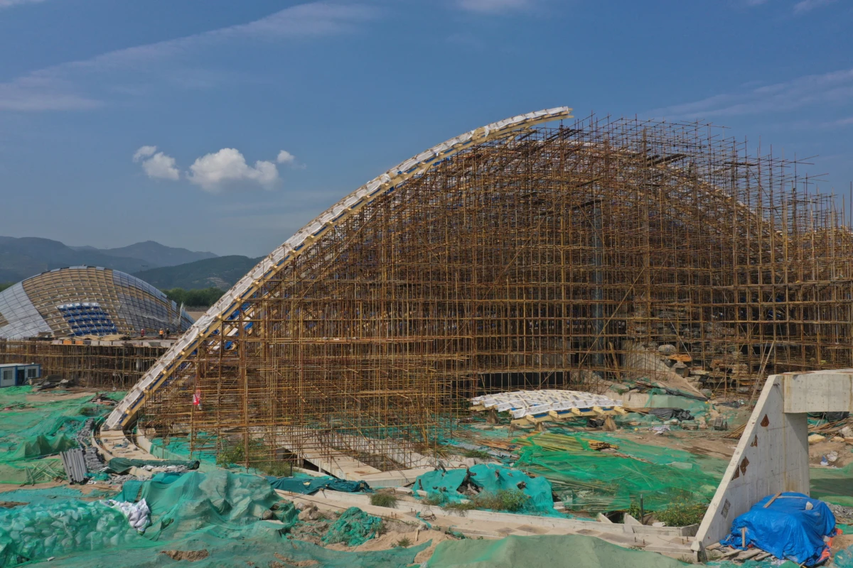 Arch-shaped structure under construction with dense bamboo scaffolding at an outdoor site; green tarps and distant hills in the background.