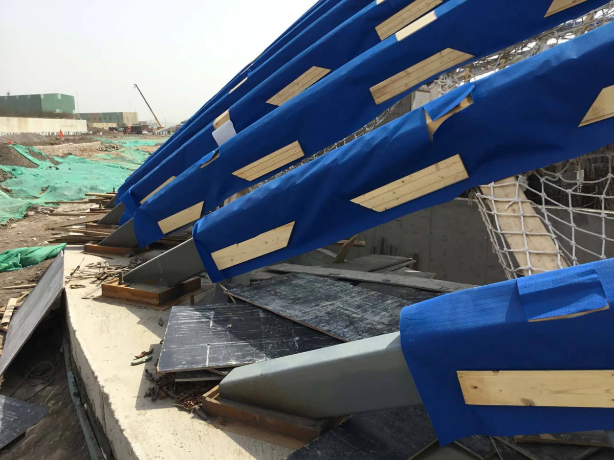 Construction site with blue protective tarps attached to curved barriers, wooden planks, and green safety netting around a concrete trench.