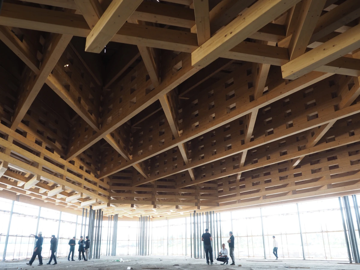 Wooden roof trusses form an open ceiling in a construction site; several workers stand and walk near large glass walls.
